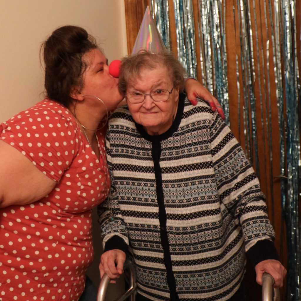 girl wearing a red and white polka dot top and a clown nose kissing an older lady wearing a birthday hat on the cheeck 